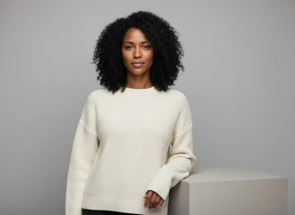 The AI model Maya wearing the ivory sweater in a grey studio, leaning on a white pedestal, generated by sameface